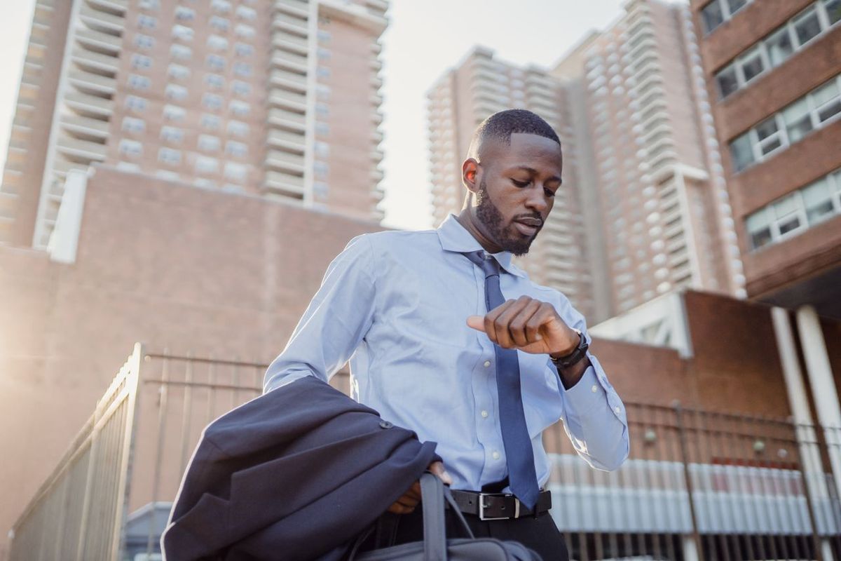 Business traveler checking a watch while carrying a suit jacket and bag in a city setting.