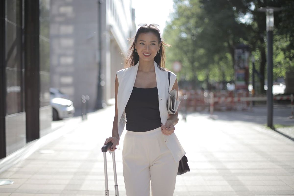 Person walking on a city sidewalk while pulling a rolling suitcase and carrying documents.