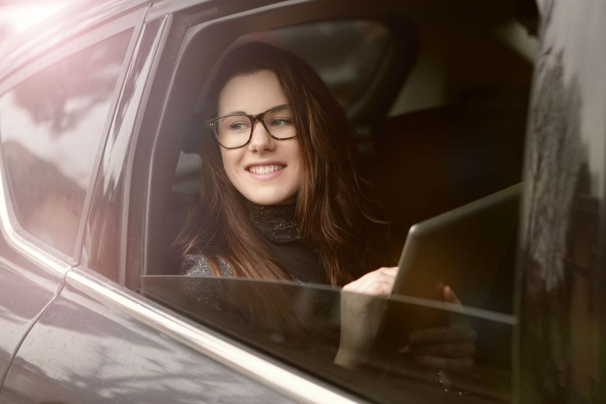 Smiling woman with glasses looking out car window, holding a tablet during daytime travel.