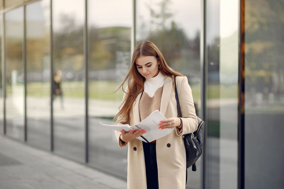 Person reading documents while standing outside a modern glass-fronted building.