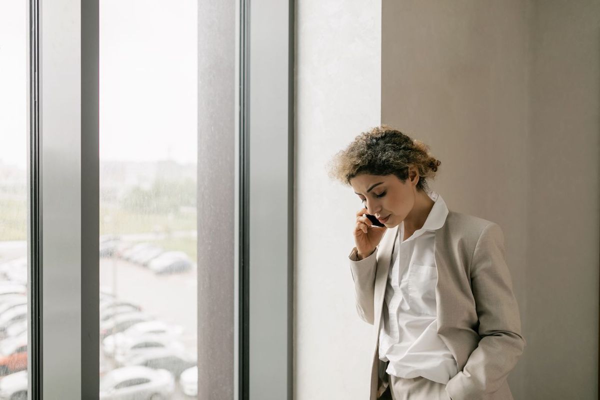 Person in a light suit talking on a smartphone while standing beside a large window.