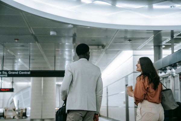 Two travelers walking through a transit terminal with carry-on luggage and coffee.