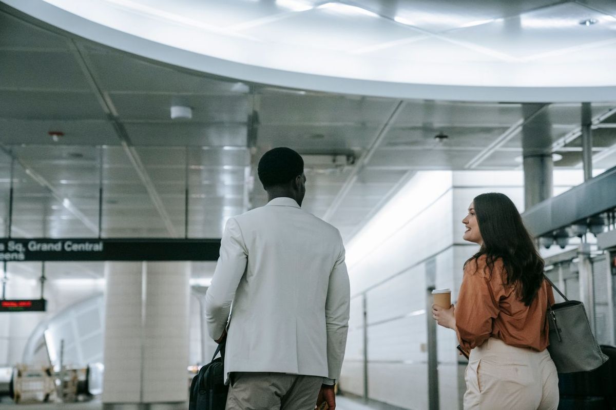 Two travelers walking through a transit terminal with carry-on luggage and coffee.
