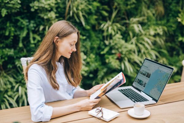 Caucasian woman reading a magazine at a wooden table with a laptop, outdoors.