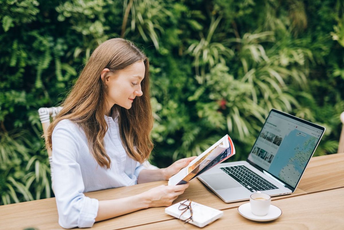 Caucasian woman reading a magazine at a wooden table with a laptop, outdoors.