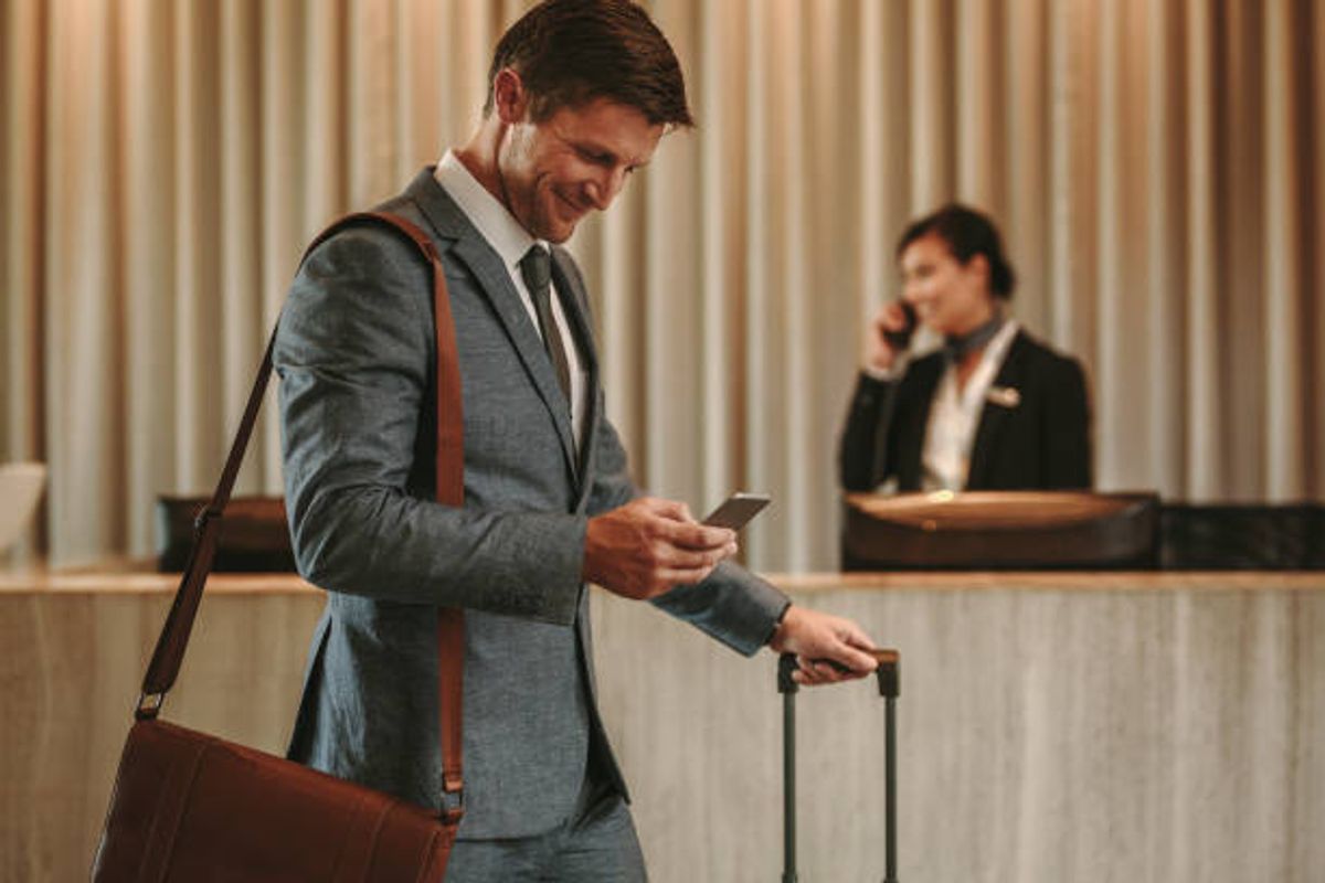 Business traveler in a grey suit checking his phone while wheeling luggage through a hotel lobby, with a front desk agent on a call in the background
