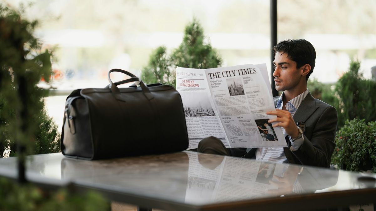 Man in suit reading newspaper outdoors with duffel bag on table.