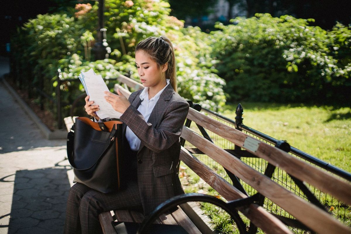 Professional sitting on a park bench reviewing documents beside a large tote bag.