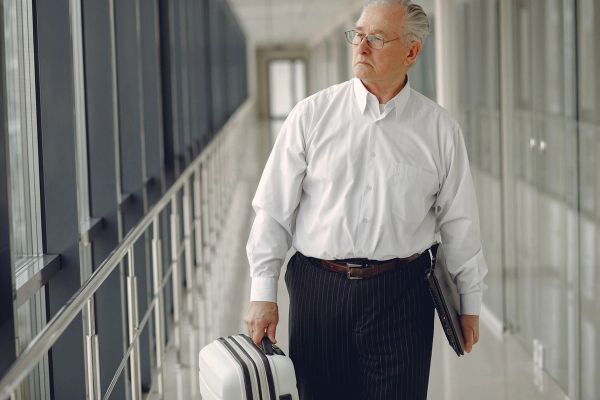 Older man in white shirt walking with suitcase and folder.
