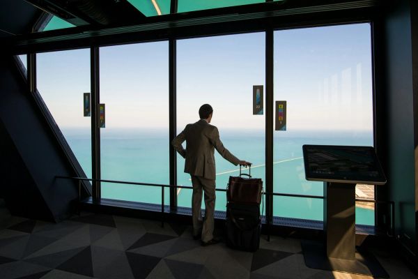 Person in suit with luggage gazing out window at water view.