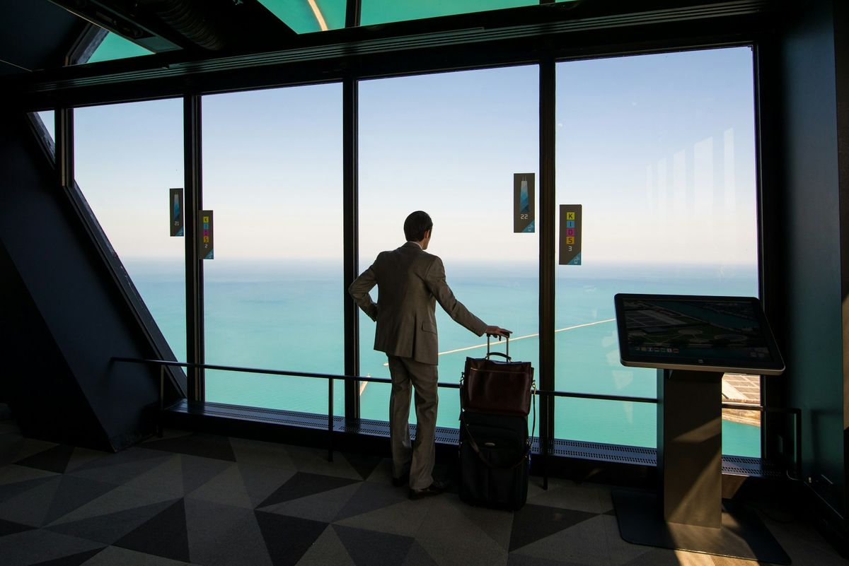 Person in suit with luggage gazing out window at water view.