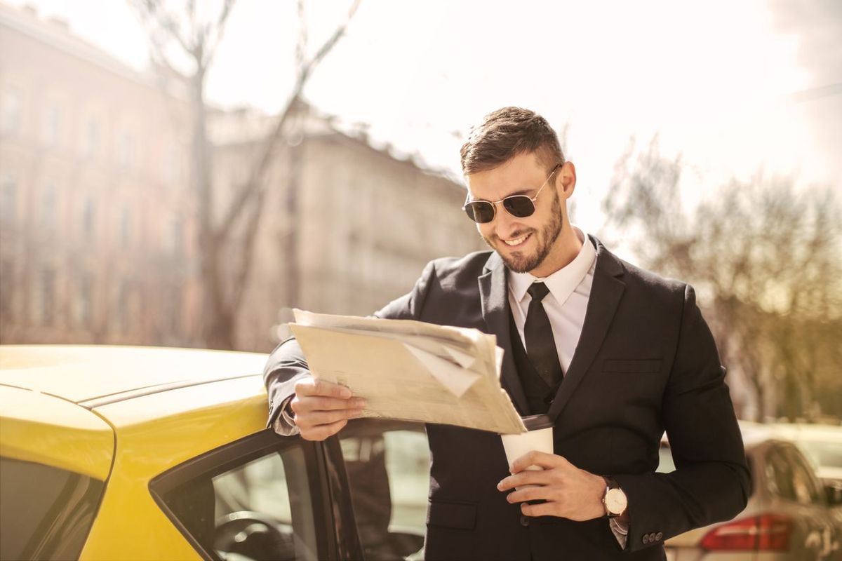 Man in suit with coffee and newspaper by car.