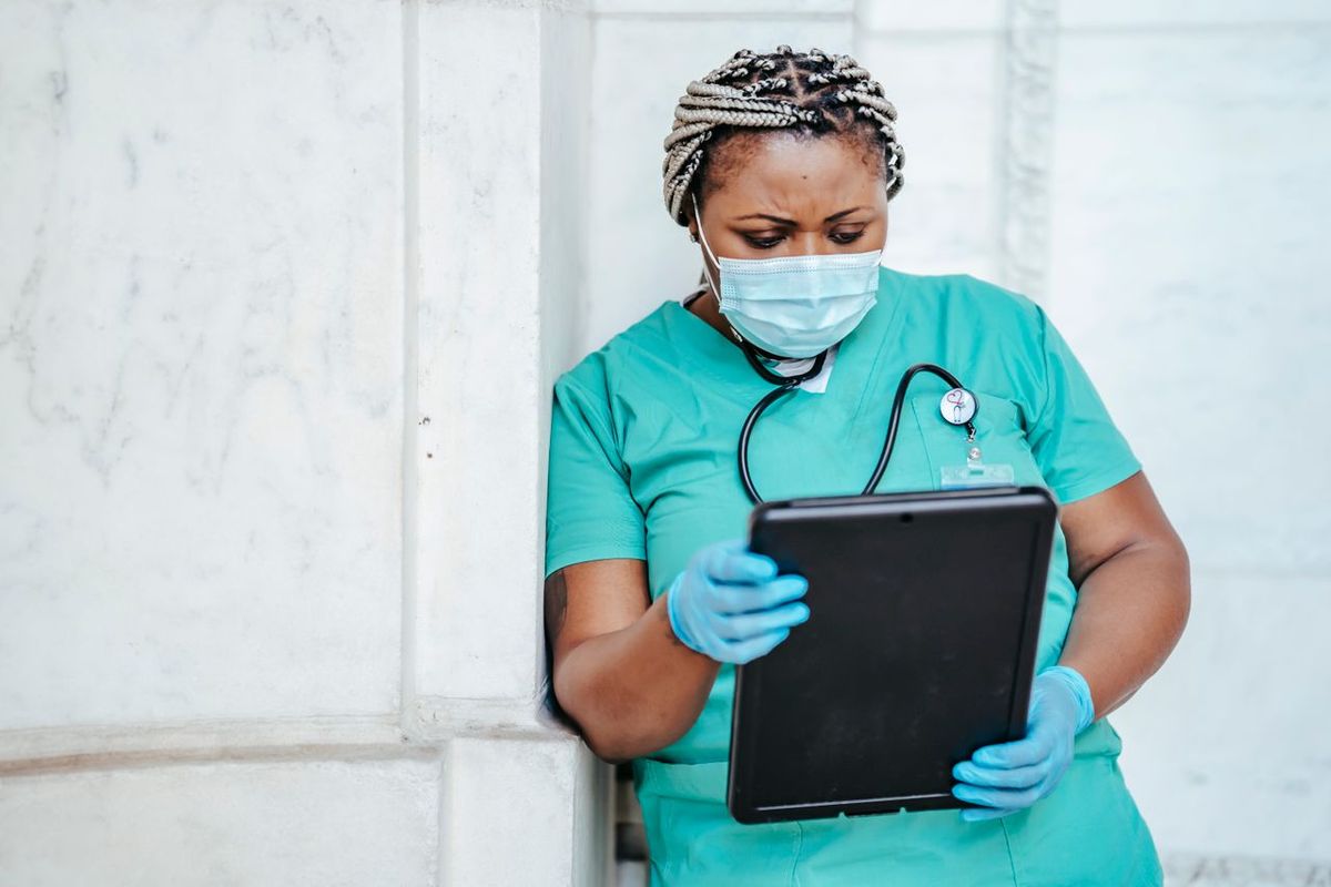 Female nurse in scrubs and mask using tablet for patient care documentation.
