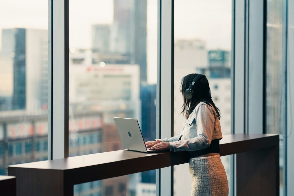 Woman wearing headphones working on laptop by window.