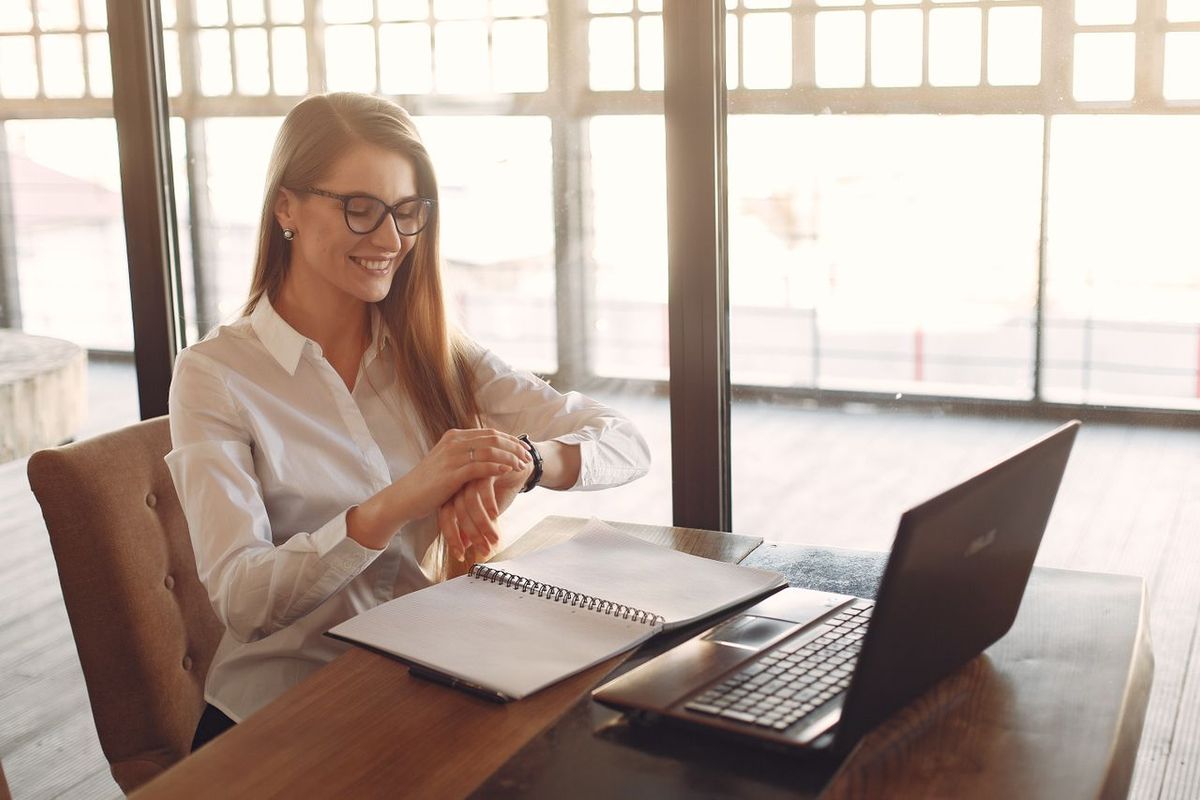 Person at a desk checking a watch beside a laptop and notebook.