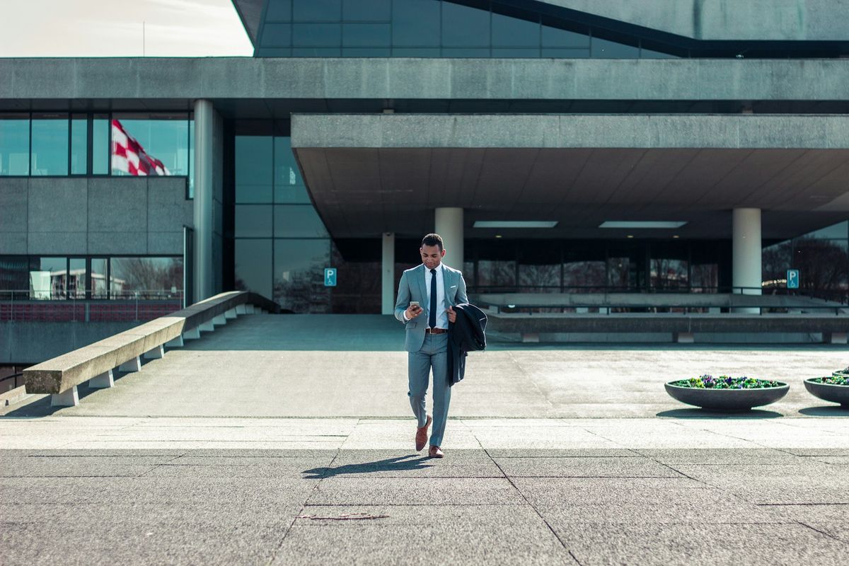 Person in gray suit walking outside modern glass building with phone and jacket.