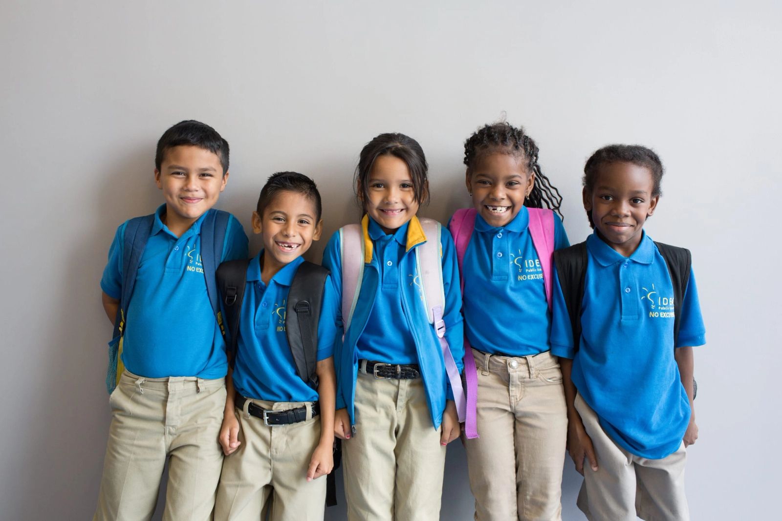 Five IDEA Public Schools students in blue uniform polos and backpacks, smiling in front of a grey wall