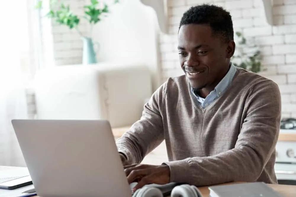 Smiling professional black business man using laptop computer working from home office. Happy african male customer makes ecommerce delivery order from supermarket typing on pc sits at kitchen table.