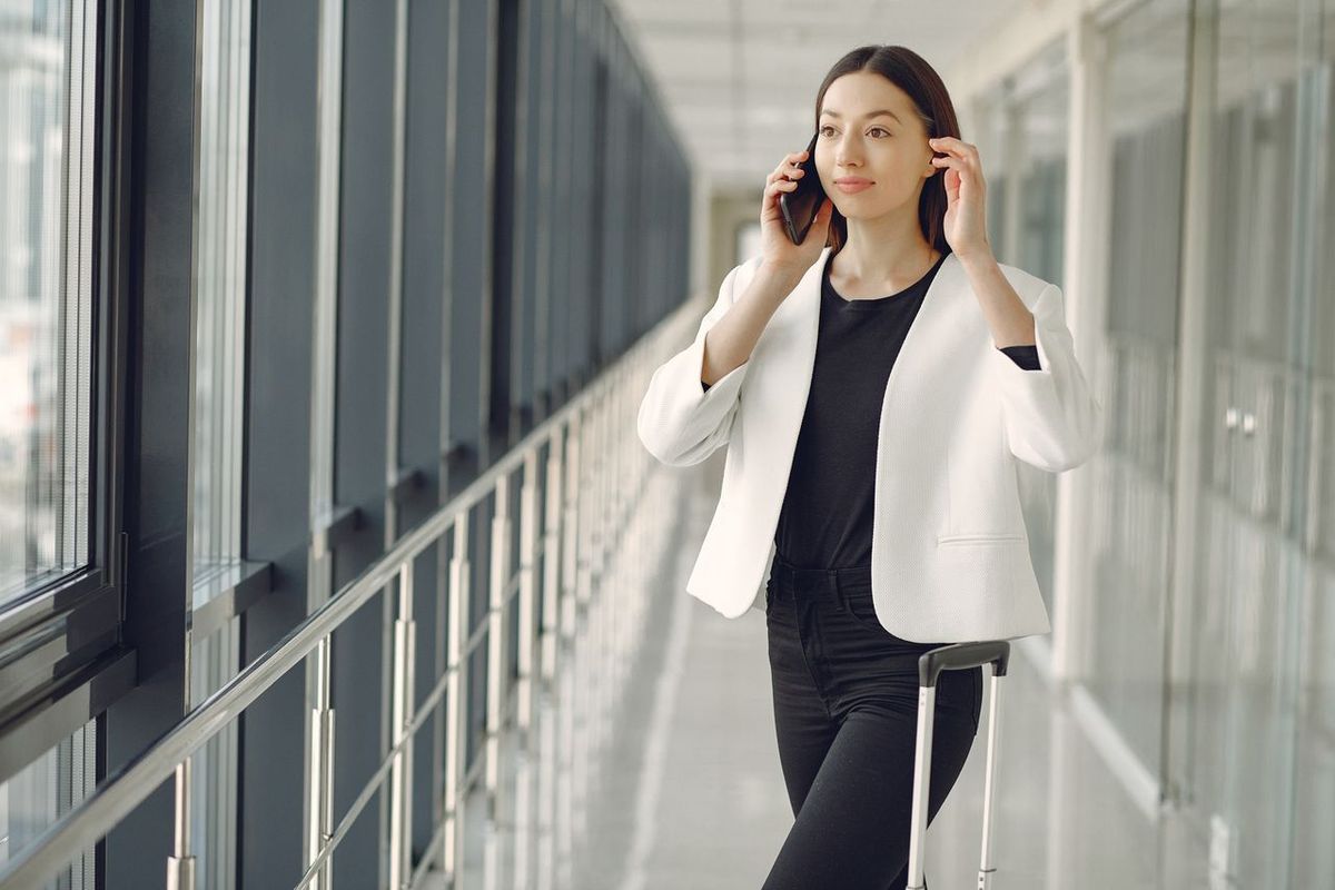 Person in a white blazer talking on a smartphone while pulling a suitcase through a bright corridor.