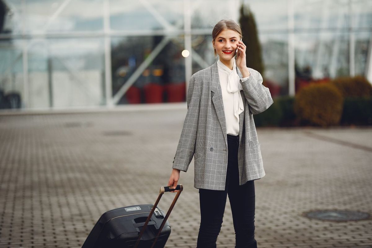 A woman in business attire talking on a phone while pulling a suitcase outdoors.