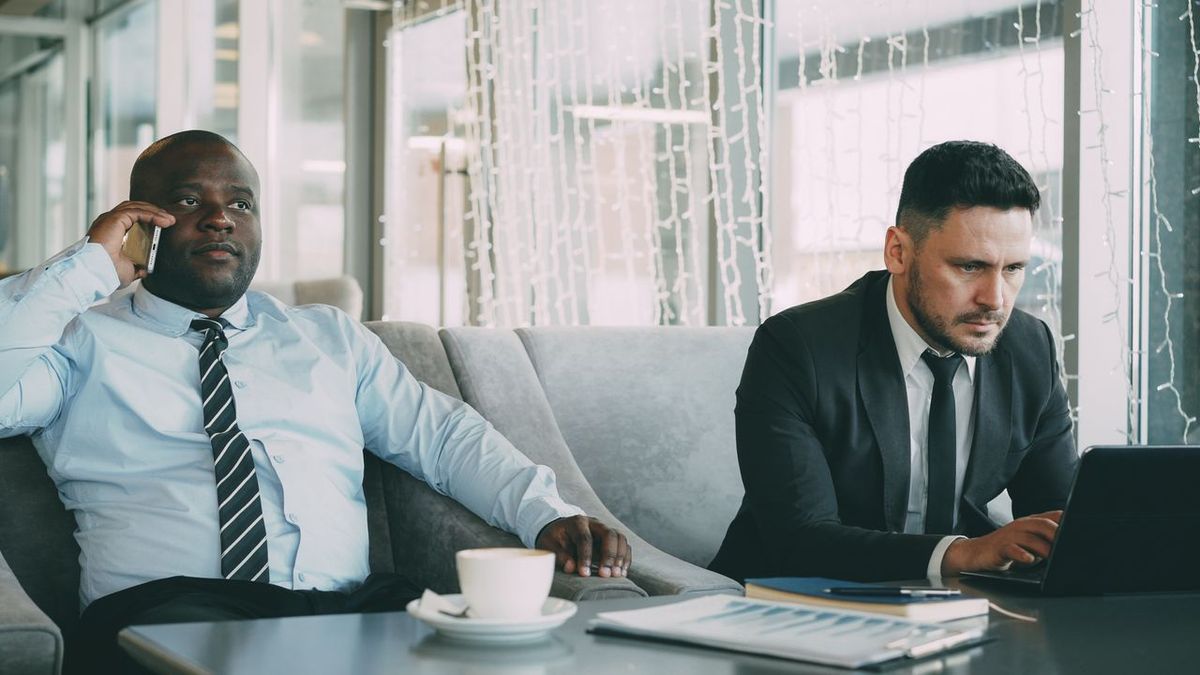 Two business professionals working in a lounge area, one on a phone call and the other using a laptop.