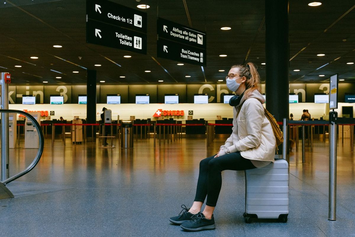 Traveler wearing a face mask seated on a suitcase in an airport check-in area.