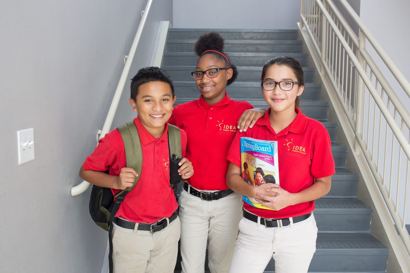 Three IDEA Public Schools students in red uniform polos on a campus stairwell, one holding a SpringBoard textbook
