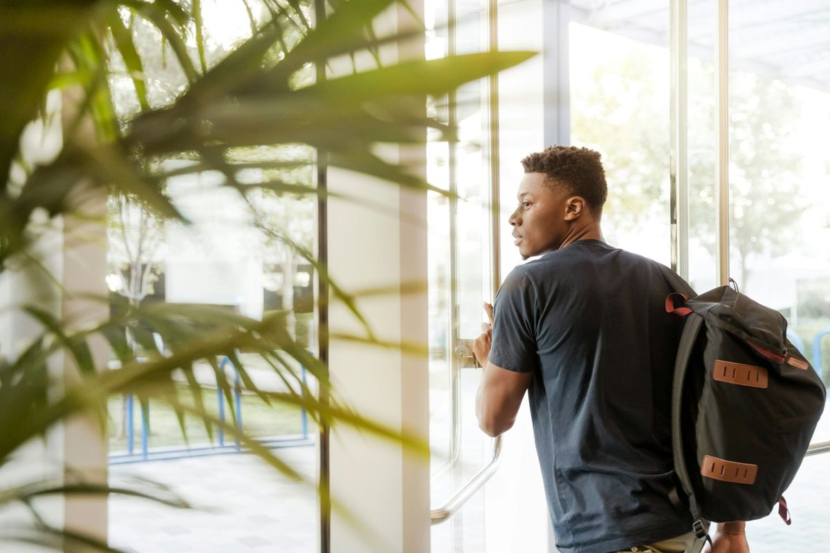 Person with a backpack opening a glass door in a bright building lobby with plants in the foreground.