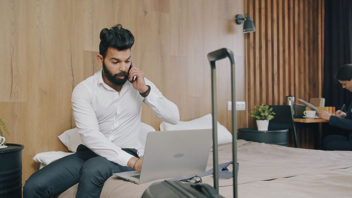 Person on bed using laptop and phone with suitcase nearby.