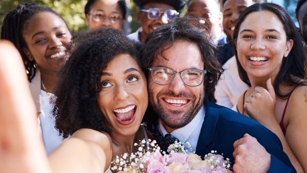Wedding couple taking a joyful selfie with friends and family gathered around them outdoors.