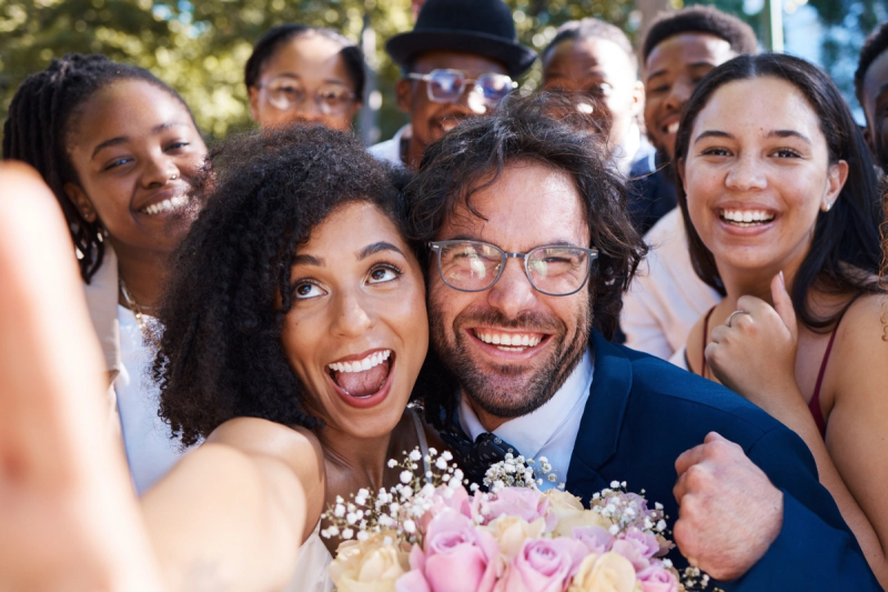 Wedding couple taking a joyful selfie with friends and family gathered around them outdoors.