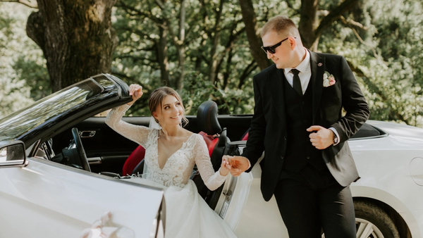 Bride stepping out of a white convertible while holding hands with the groom in an outdoor wooded setting.