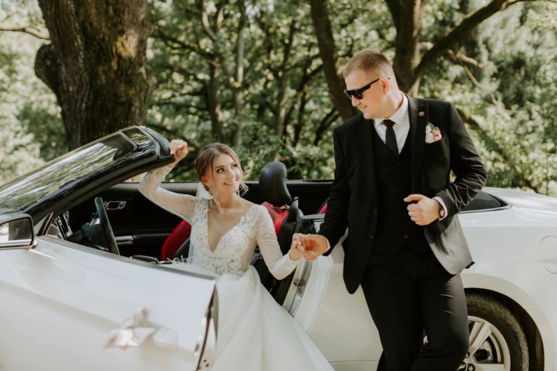 Bride stepping out of a white convertible while holding hands with the groom in an outdoor wooded setting.