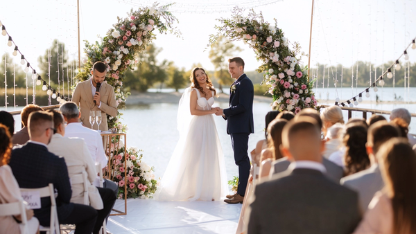 Bride and groom holding hands during outdoor lakeside wedding ceremony under floral arch, with officiant and seated guests