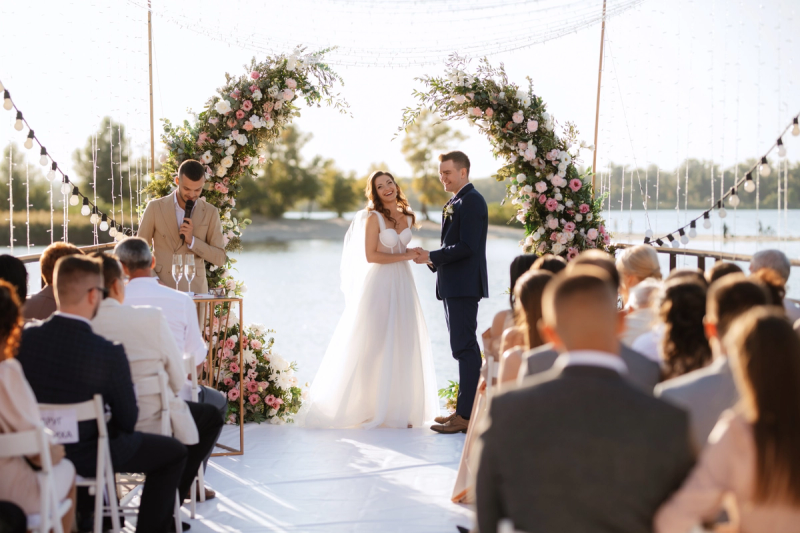 Bride and groom holding hands during outdoor lakeside wedding ceremony under floral arch, with officiant and seated guests