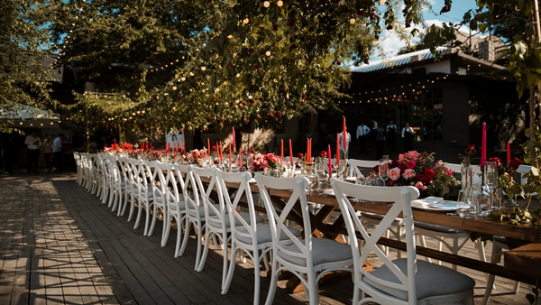 Outdoor wedding reception with a long wooden banquet table, white chairs, red and pink floral arrangements, tall candles, and string lights surrounded by greenery.