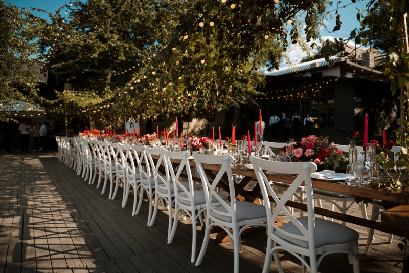 Outdoor wedding reception with a long wooden banquet table, white chairs, red and pink floral arrangements, tall candles, and string lights surrounded by greenery.