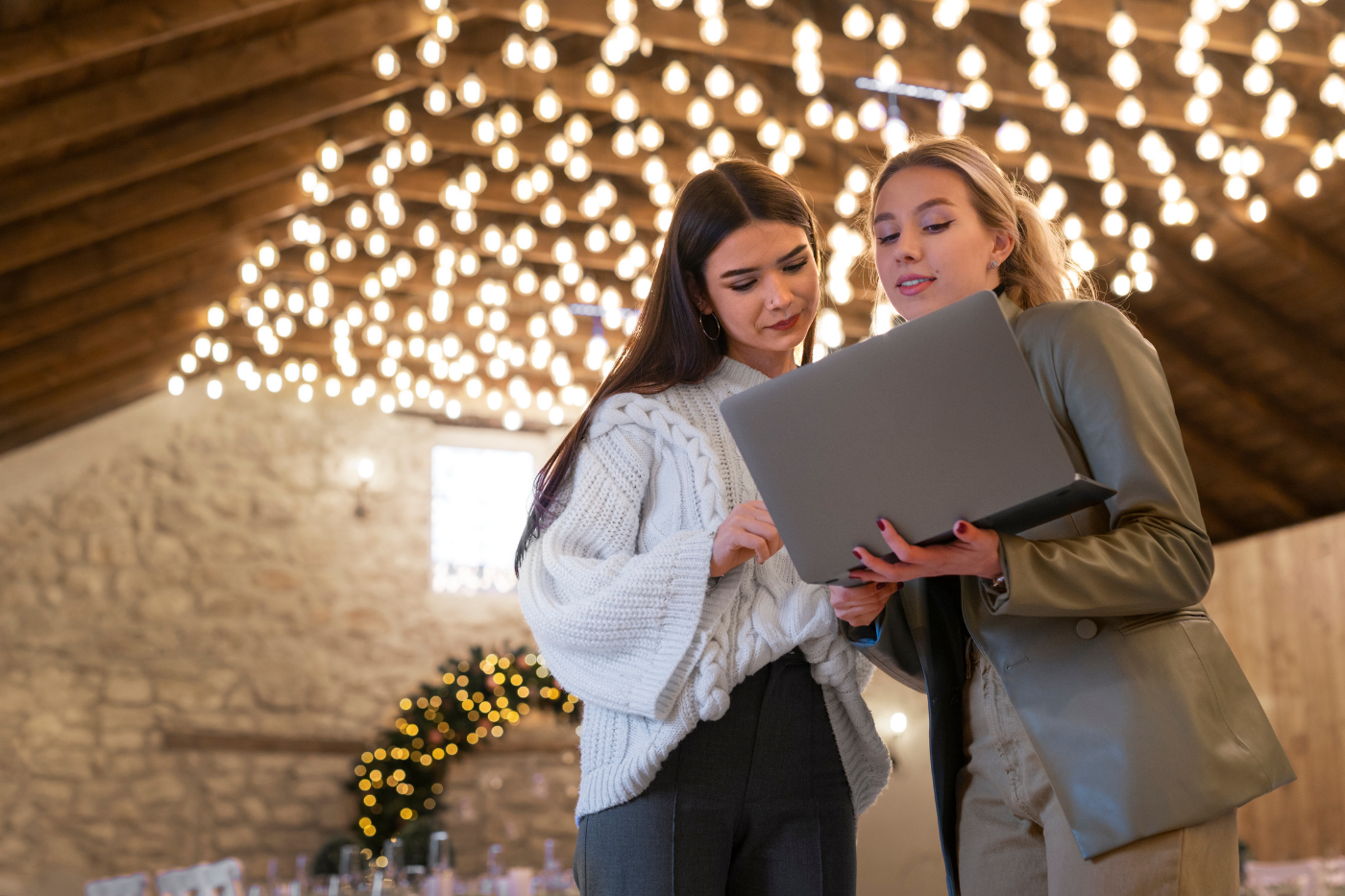 Two women reviewing a laptop together inside a warmly lit event venue decorated with hanging string lights.