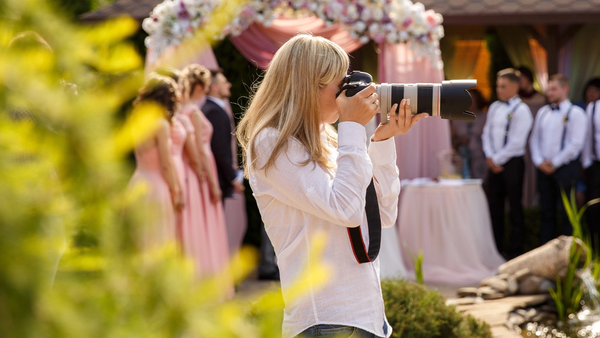 Wedding photographer taking photos during an outdoor ceremony, with the wedding party and a floral arch in the background.