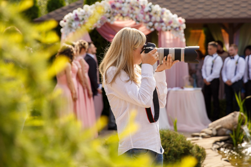 Wedding photographer taking photos during an outdoor ceremony, with the wedding party and a floral arch in the background.