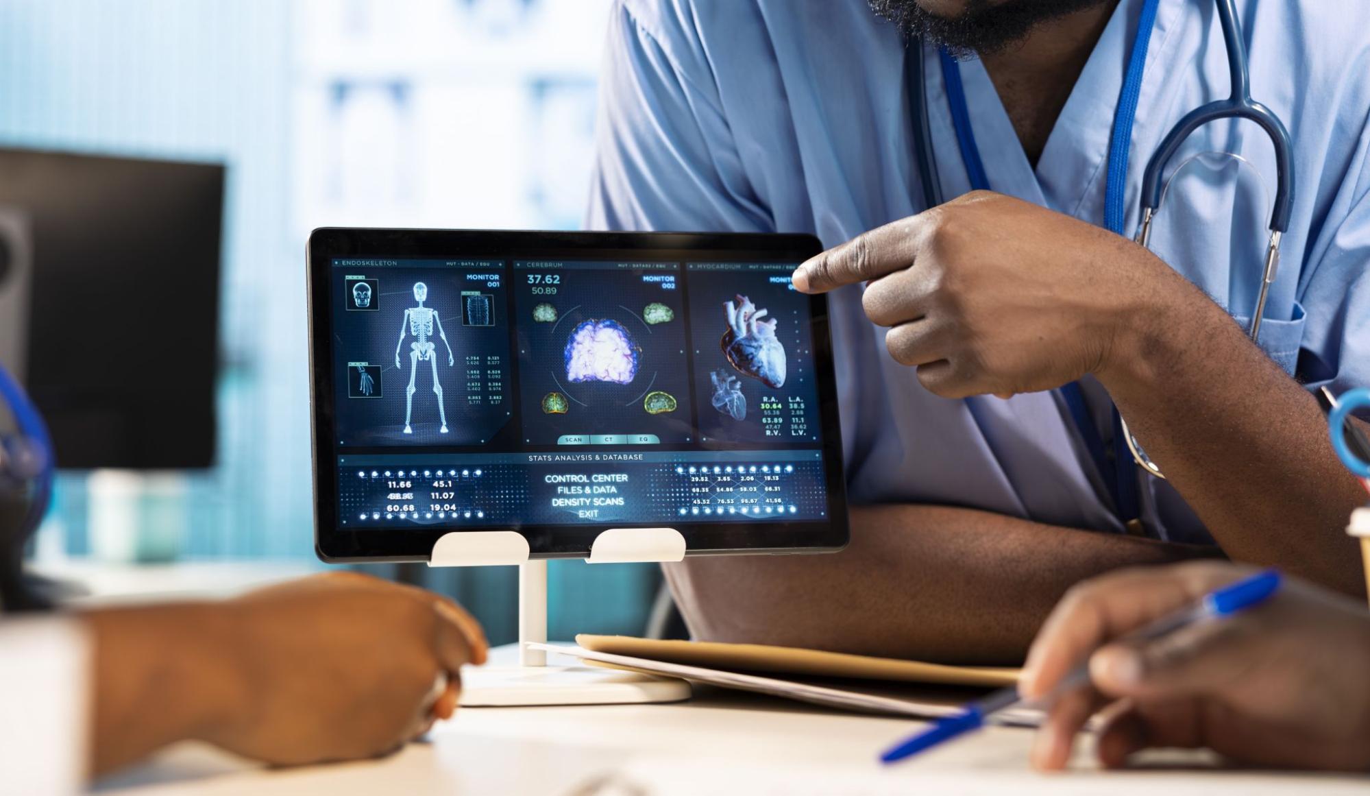 A doctor points to a tablet screen, discussing information with a patient in a clinical setting.
