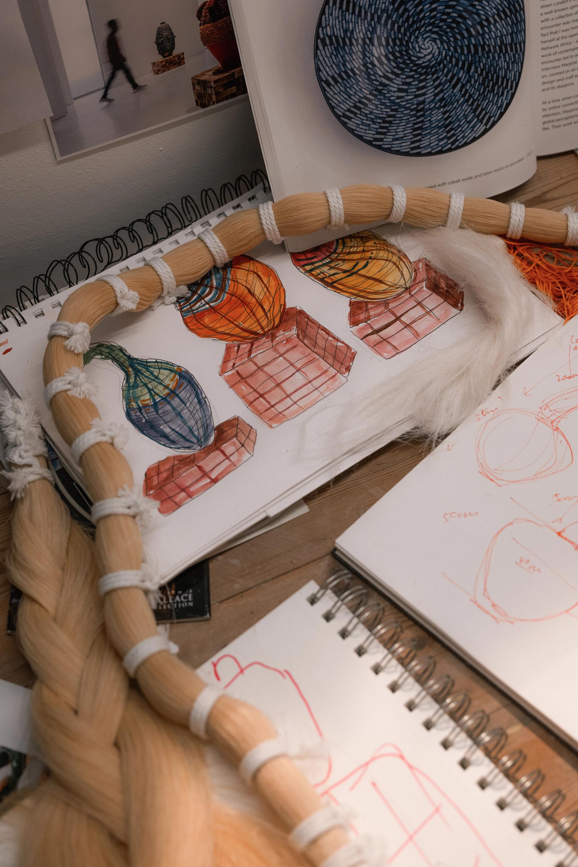 A studio desk with sketchbooks showing colorful designs for woven vessel sculptures, alongside a braided fiber material sample and reference books.