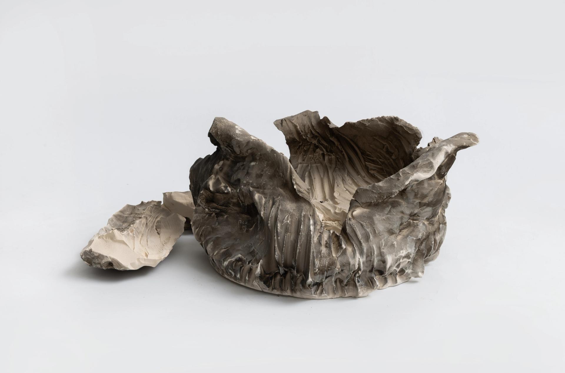 Rough-hewn gray ceramic vessel with a fractured rim, displayed beside a broken fragment on a white background.