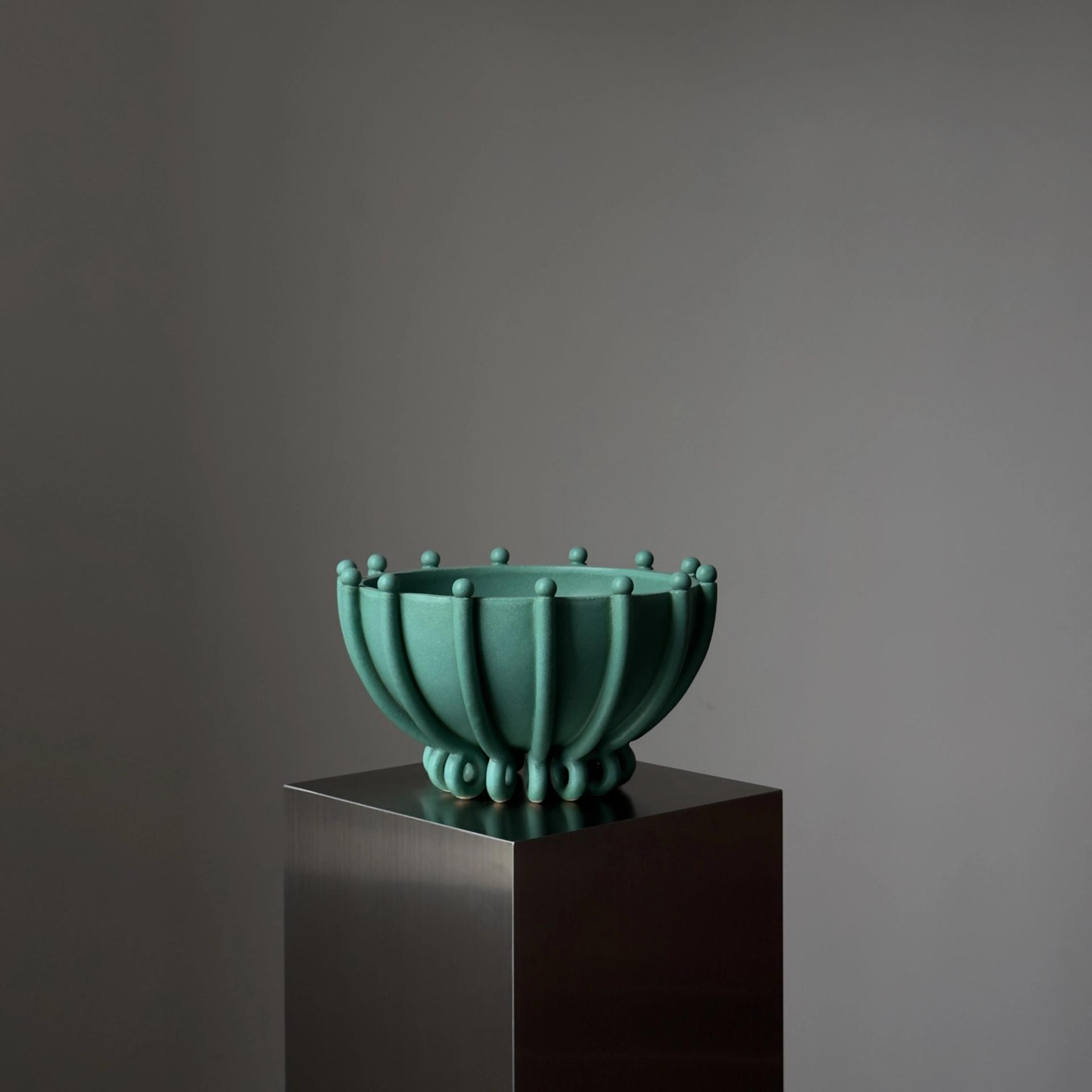 Green ceramic bowl on a dark gray plinth against a pale gray backdrop.