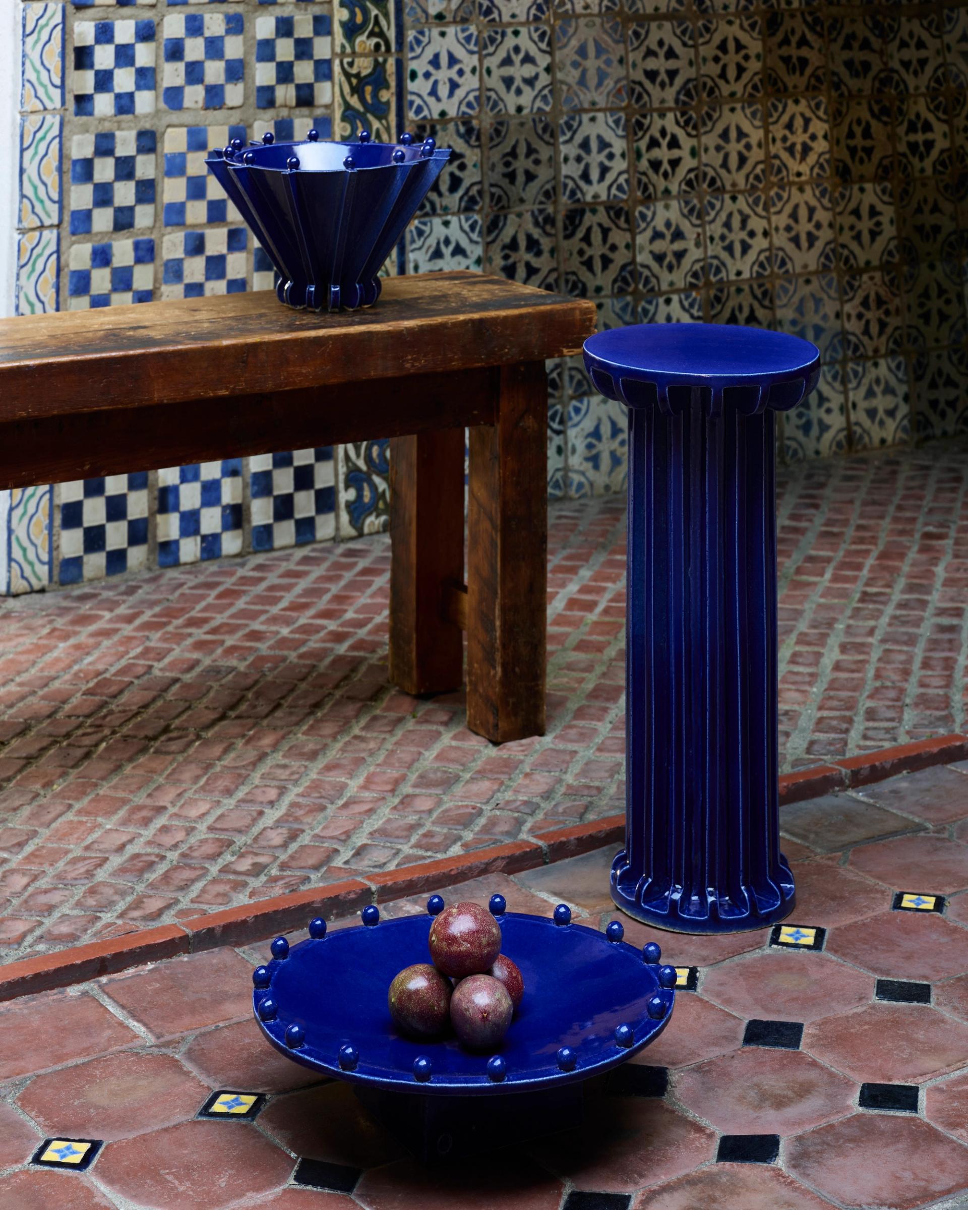 Blue ceramic vessels and pedestal displayed beside wooden table on patterned tiled floor.