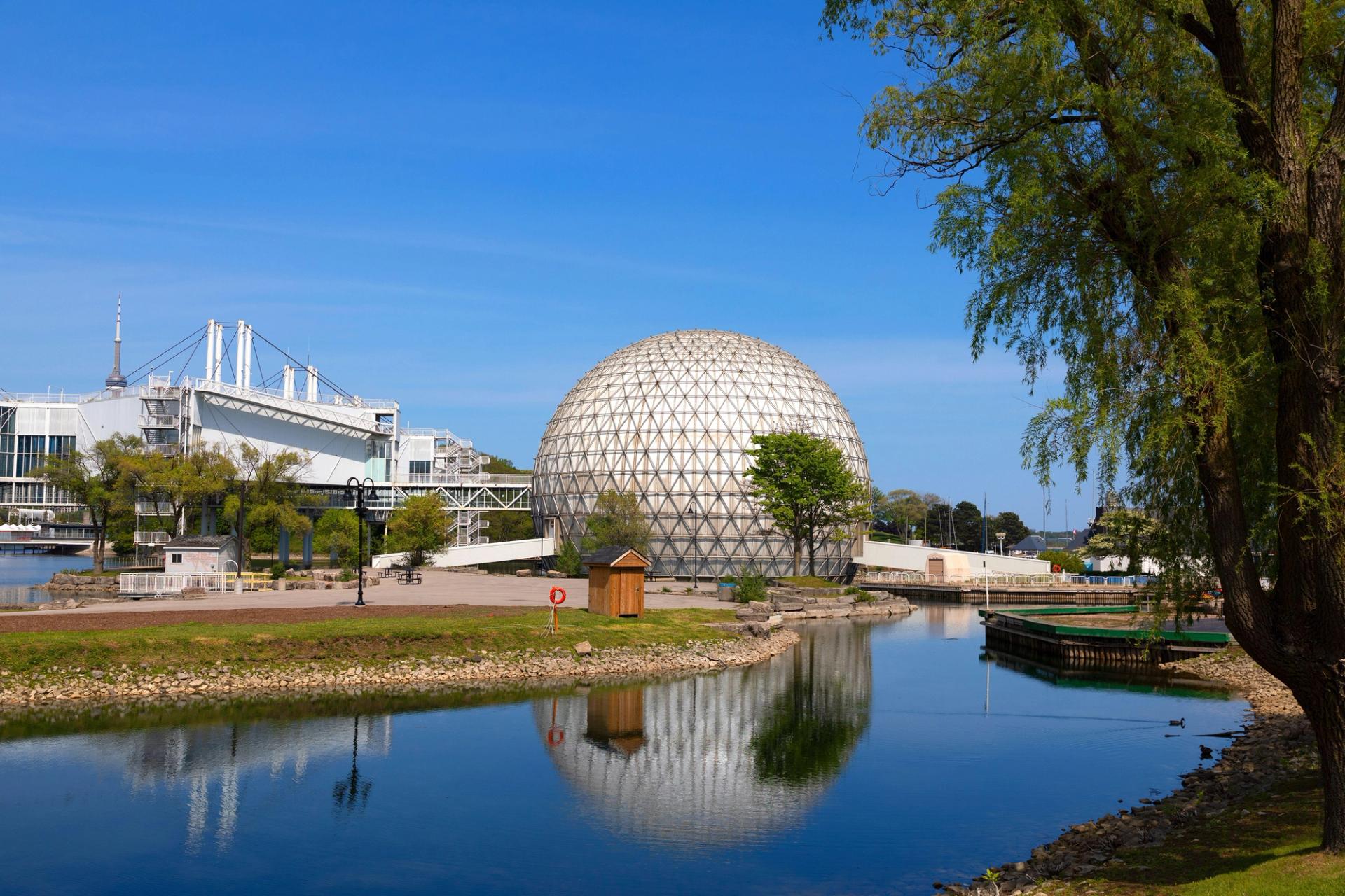 A large geodesic dome structure reflected in a calm waterway, surrounded by parkland and modern buildings under a clear blue sky