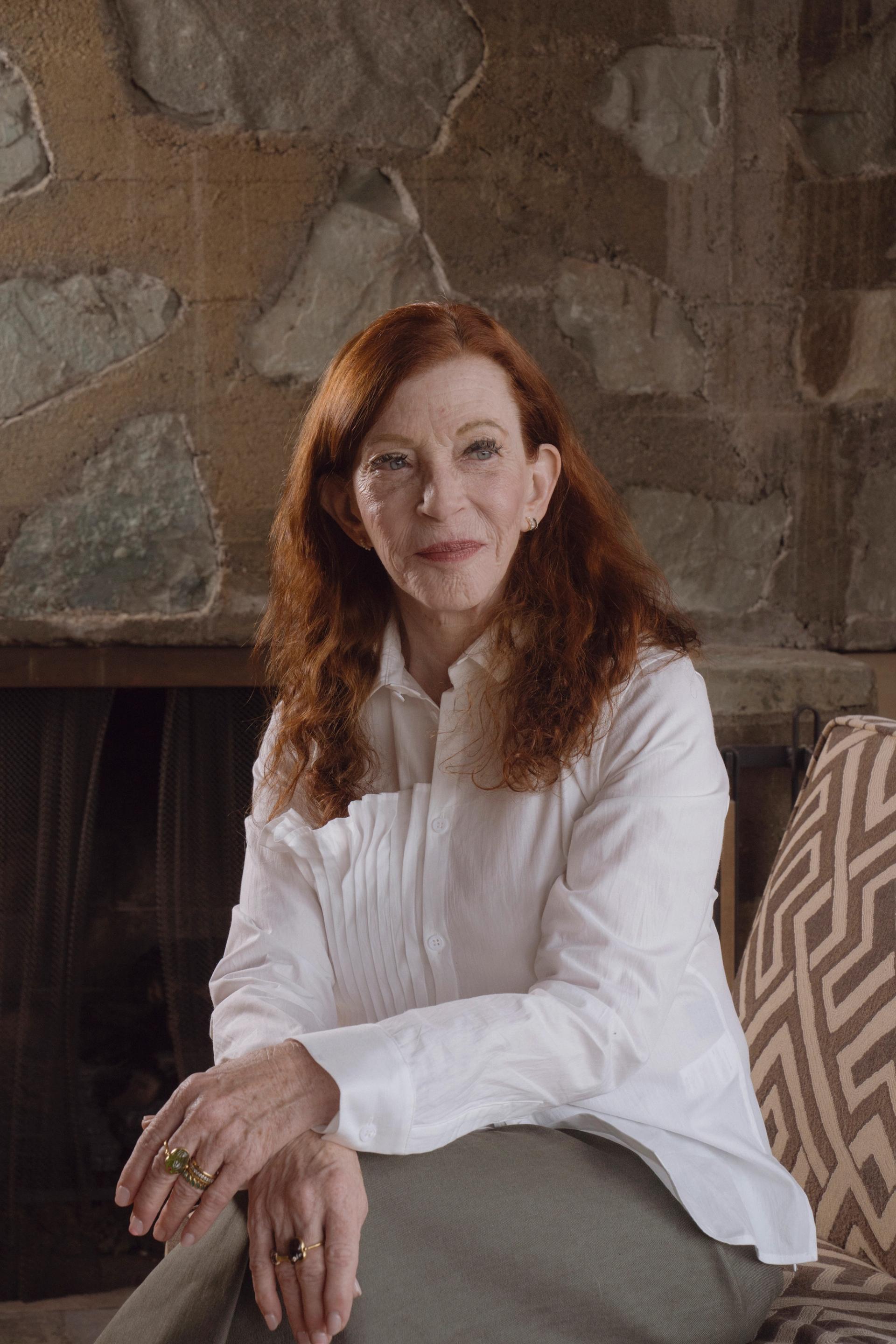 A woman with red hair sits on a chair with her hands crossed in her lap in front of a textured stone wall.