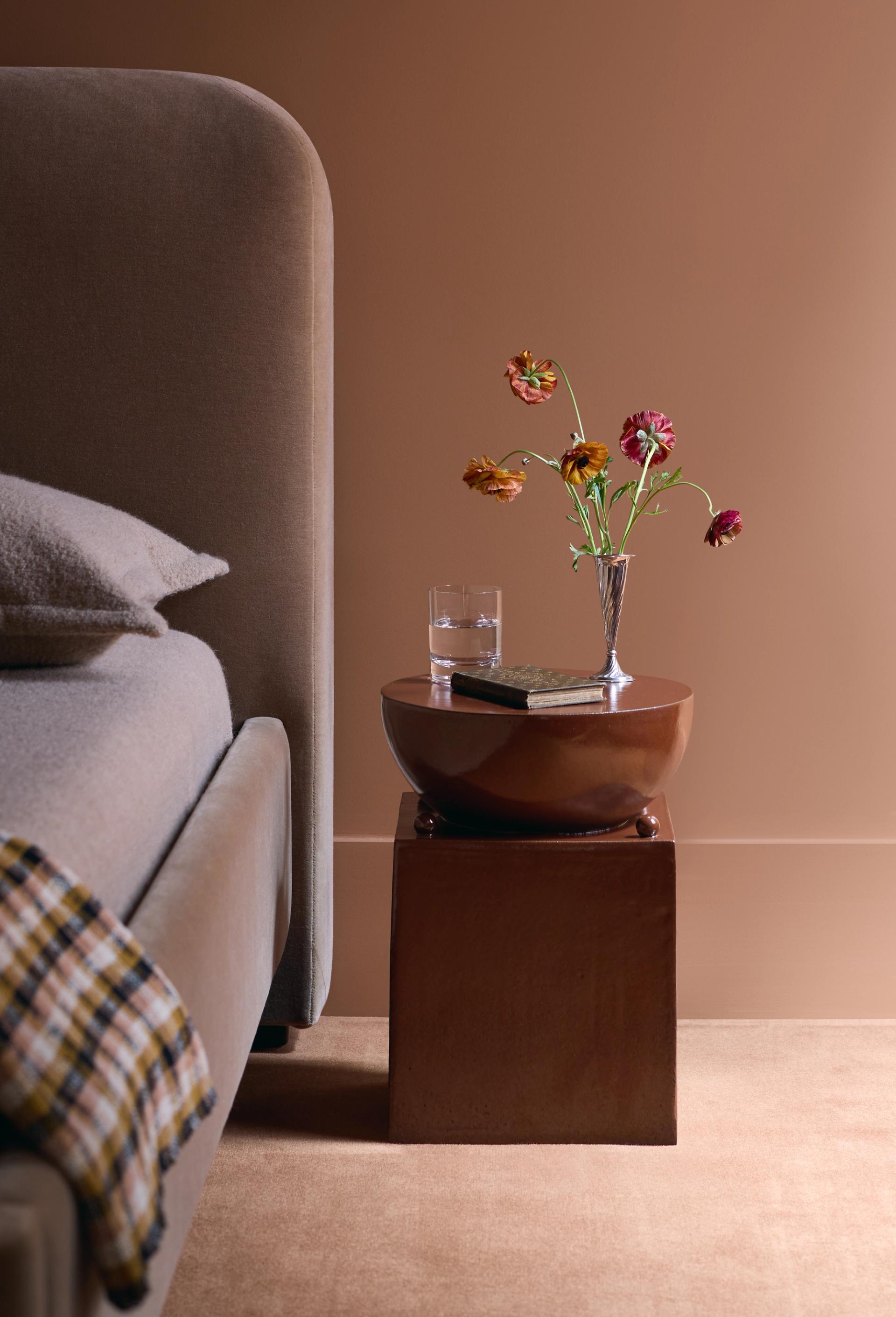 Brown ceramic bedside table with flowers, glass of water, and book beside upholstered bed against warm-toned wall.