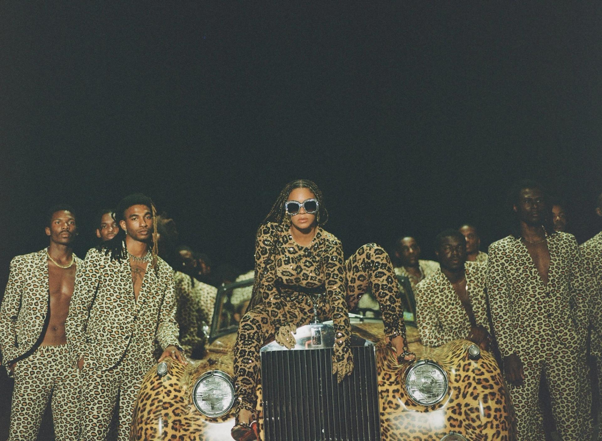 A woman in a leopard-print outfit and crystal sunglasses perches on a leopard-print car, surrounded by a group of men in matching leopard-print suits, against a dark background.
