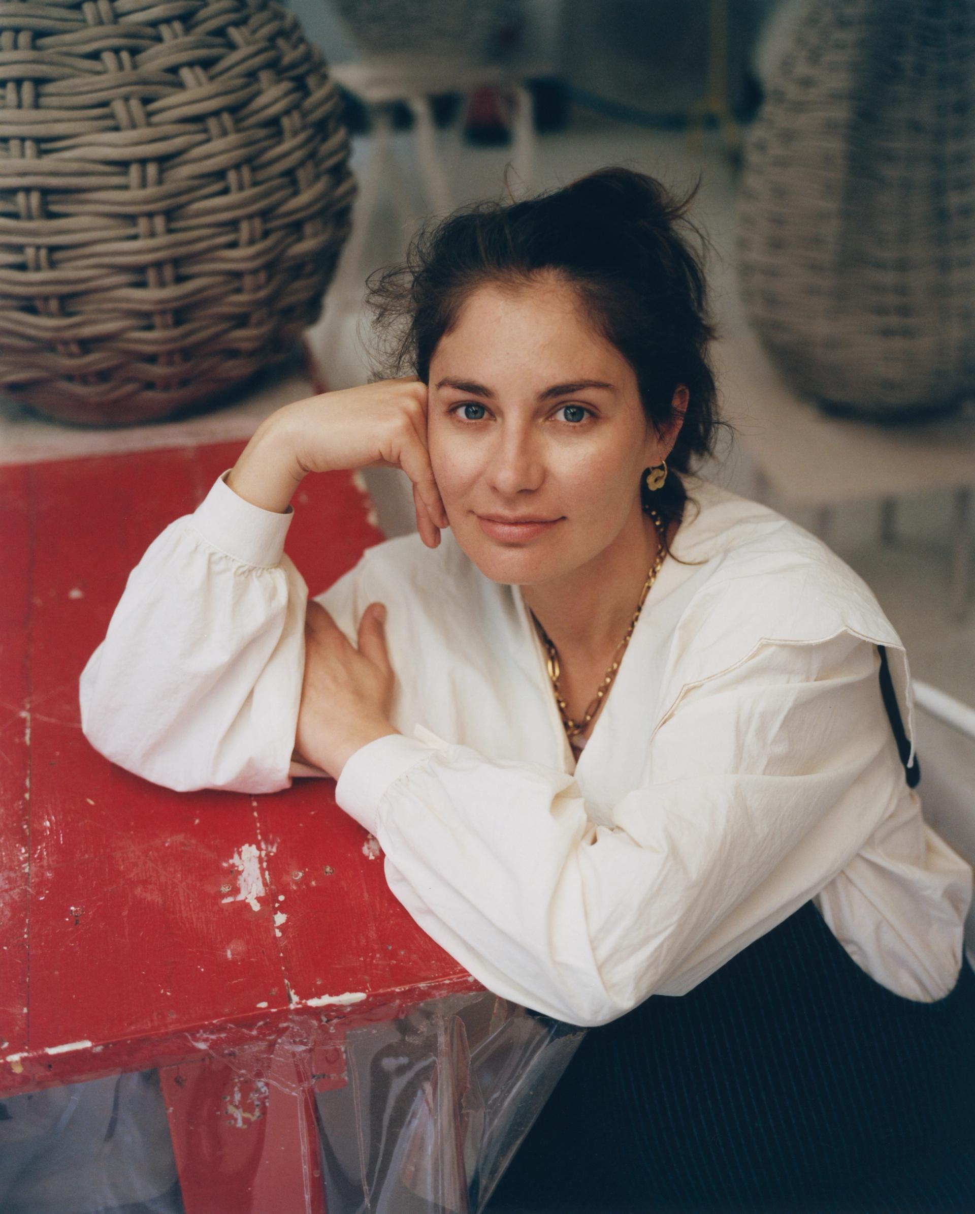 A woman in a white blouse and gold jewelry rests her arms on a worn red surface, with large woven basket sculptures visible behind her.